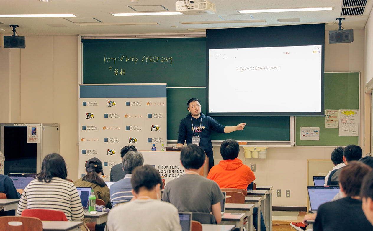 写真:ワークショップの様子。黒板を背に講師の井関さんが教卓に立って説明している。参加者は教室の各席にてノートPCを開き説明を受けている。